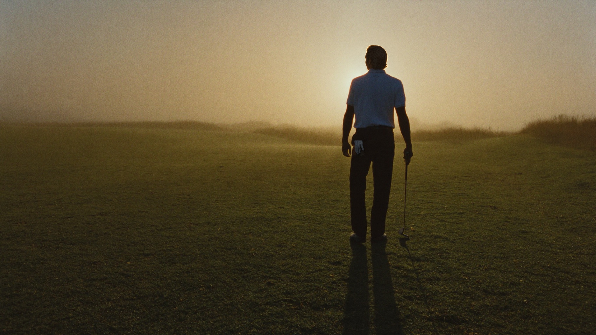 Een golfer op de rug gezien, met een ijzer in de hand, kijkt over een fairway in de ochtendnevel