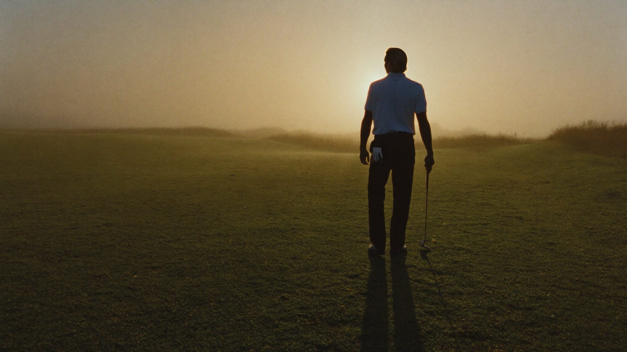Een golfer op de rug gezien, alleen op de fairway in de ochtendnevel, de zon recht voor hem